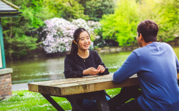 Two students sat at a picnic bench in a park, chatting and smiling together